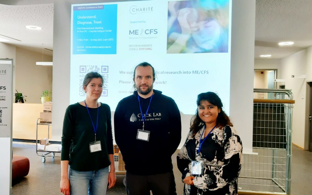 Drei Forschende, die einen Posterpreis gewonnen haben stehen vor einer Powerpointpräsentation auf der internationalen Fachtagung zu ME/CFS an der Charité Berlin: Charlotte Kröger (Platz 2), Martin Kräter (Platz 1) und Kanchan Dulal (Platz 3)